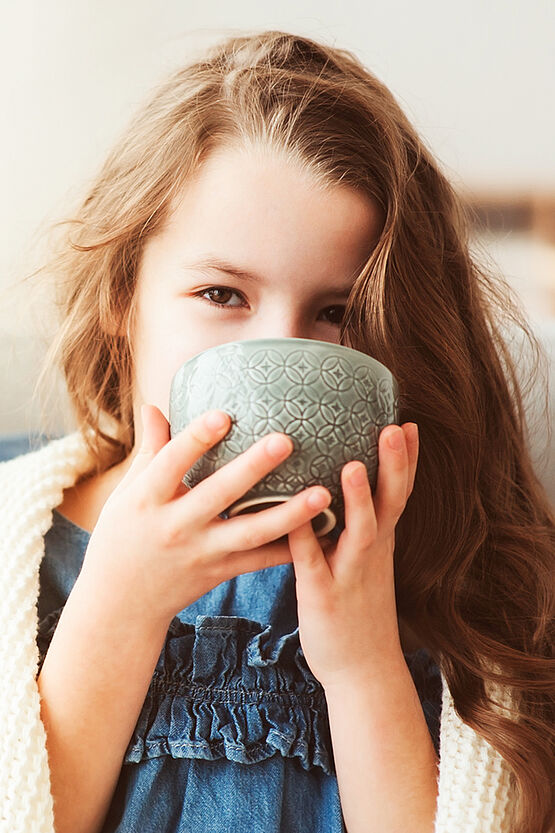 A young girl with long brown hair is holding a green cup close to her face, smiling with her eyes visible above the rim. She is wearing a blue dress and a cream-colored cardigan