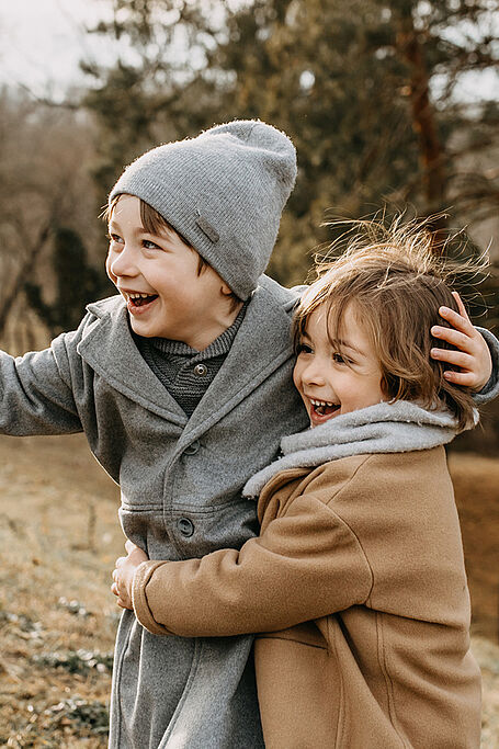 Two young children, both dressed warmly in coats and hats, joyfully embracing and laughing outdoors in a natural setting