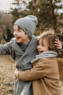 Two young children, both dressed warmly in coats and hats, joyfully embracing and laughing outdoors in a natural setting