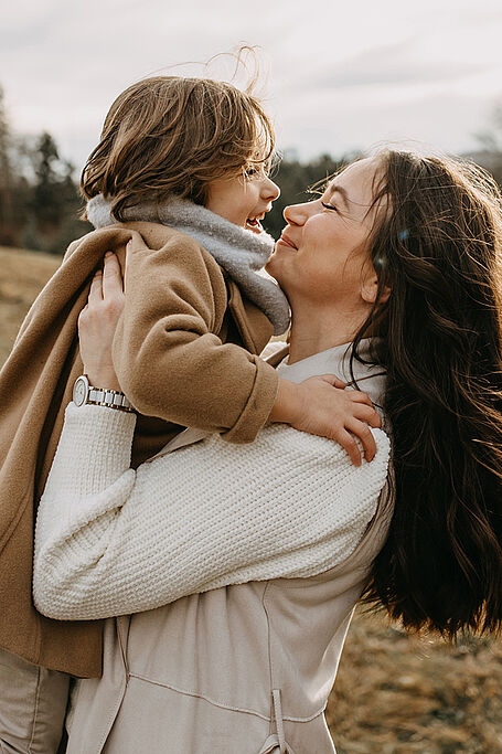 Mother holding and smiling at her child in an outdoor setting, both dressed warmly in coats and scarves on a cloudy day